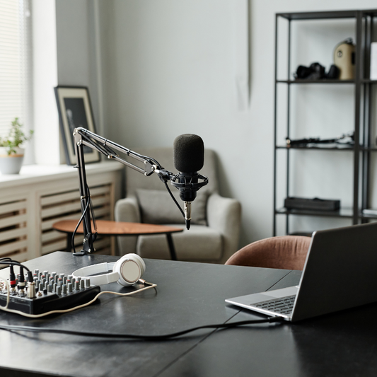 A microphone with a stand is set up on a desk, next to a laptop, keyboard, and a pair of headphones, indicating a setup for podcast production.